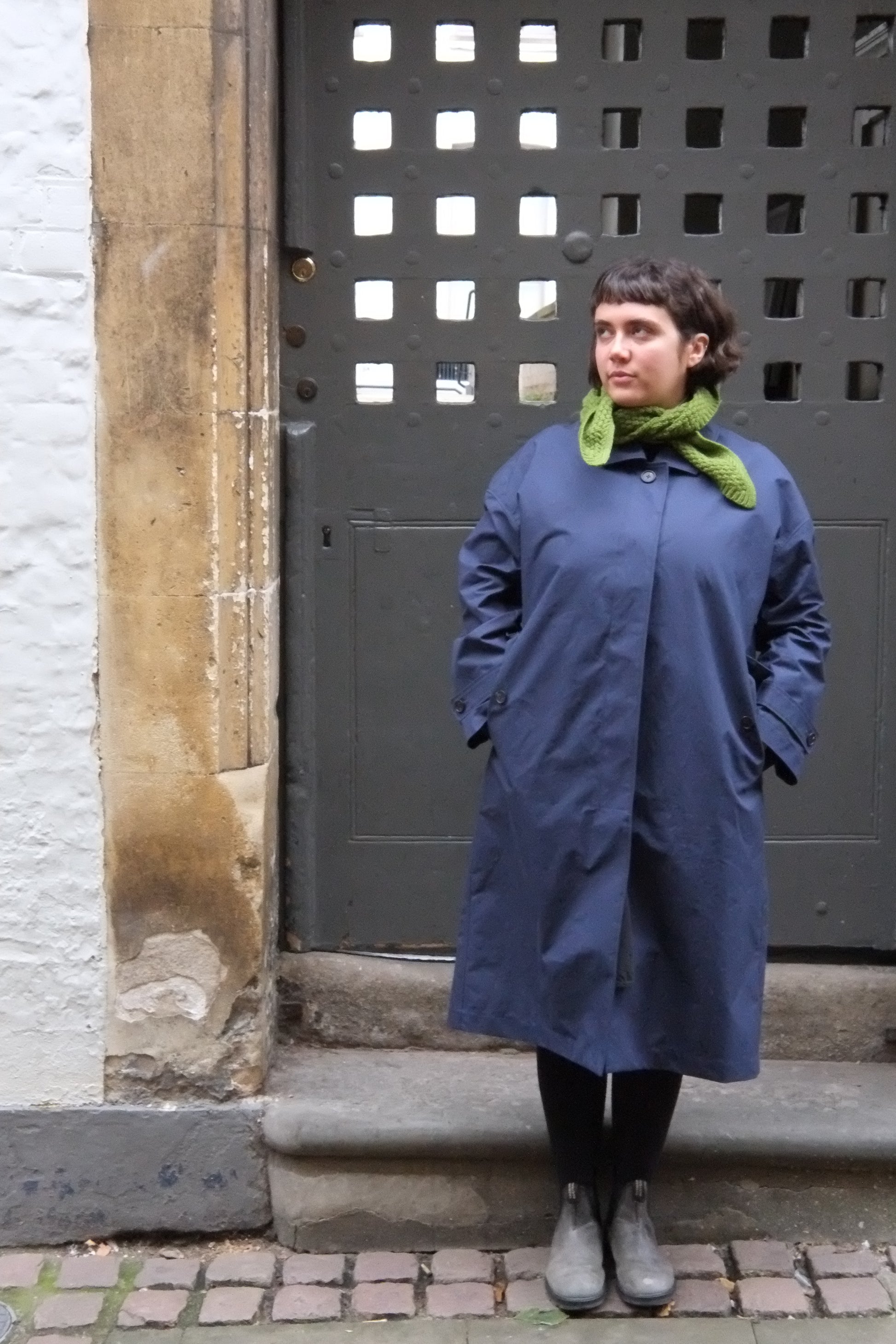 Person wearing a blue coat and green scarf standing on a sidewalk.