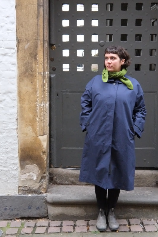 Person wearing a blue coat and green scarf standing on a sidewalk.