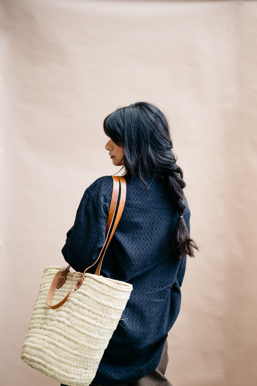 Person wearing a black textured loose fitting oversized shirt with a straw bag on a beige background