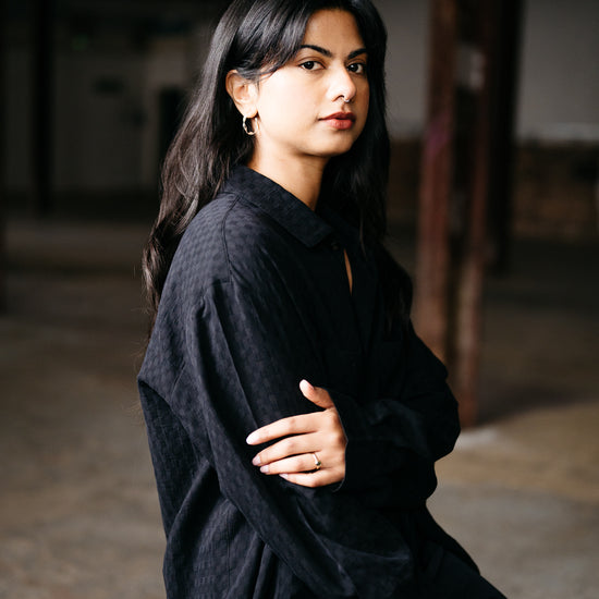 Woman wearing a black cotton textured shirt sitting on a wooden stool in a dimly lit room.