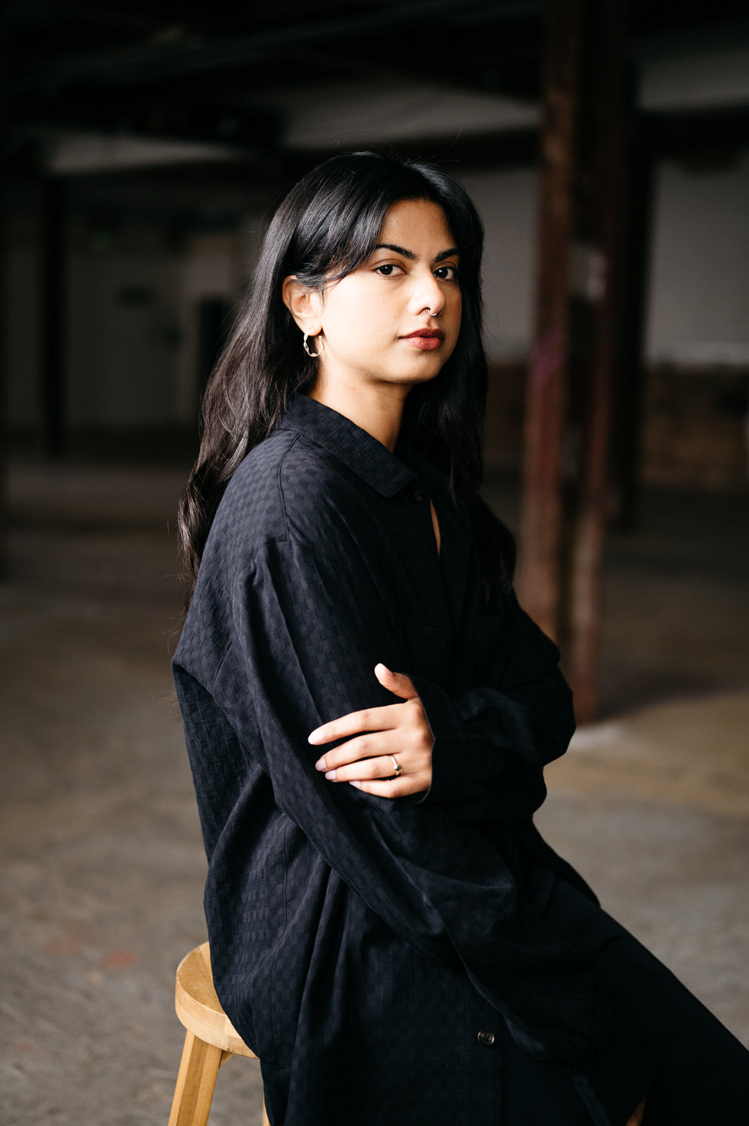 Woman wearing a black cotton textured shirt sitting on a wooden stool in a dimly lit room.
