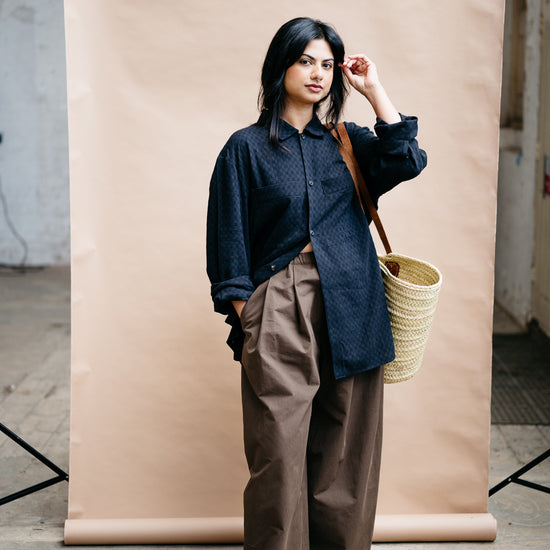 Woman in a black textured oversized shirt and brown trousers holding a woven basket against a beige backdrop with red shoes on