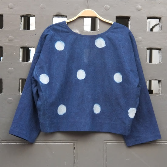 An indigo blue long-sleeve top with white polka dots, hanging on a wooden hanger against a grey grid door.