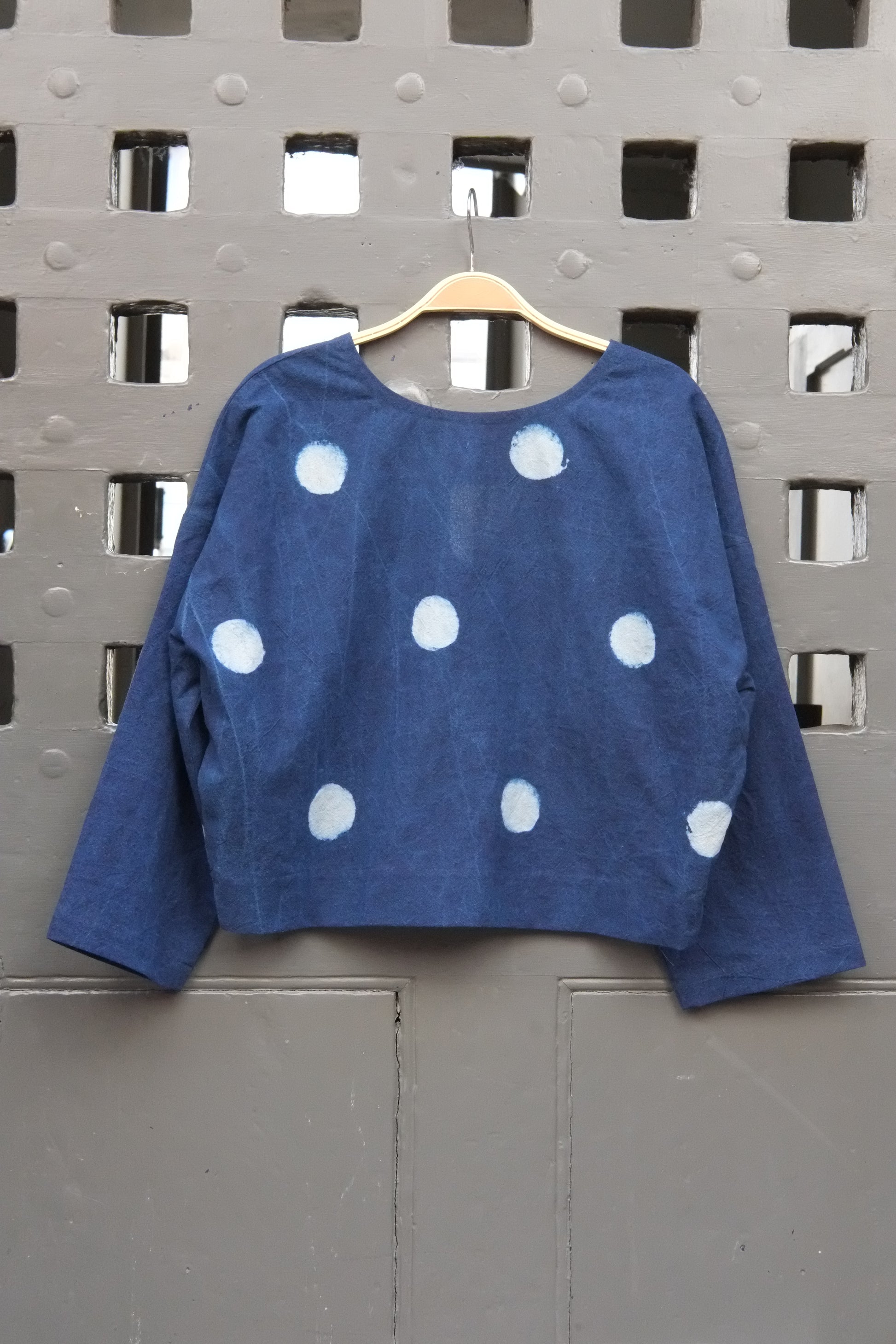 An indigo blue long-sleeve top with white polka dots, hanging on a wooden hanger against a grey grid door.