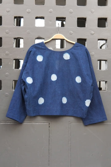 An indigo blue long-sleeve top with white polka dots, hanging on a wooden hanger against a grey grid door.