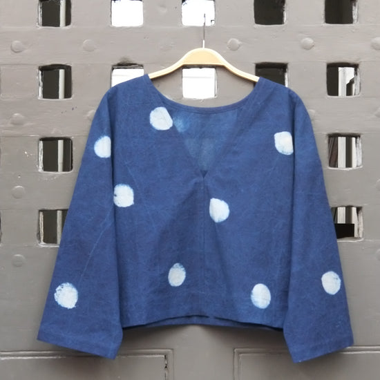 An indigo blue long-sleeve top with white polka dots hanging on a wooden hanger against a grey grid door