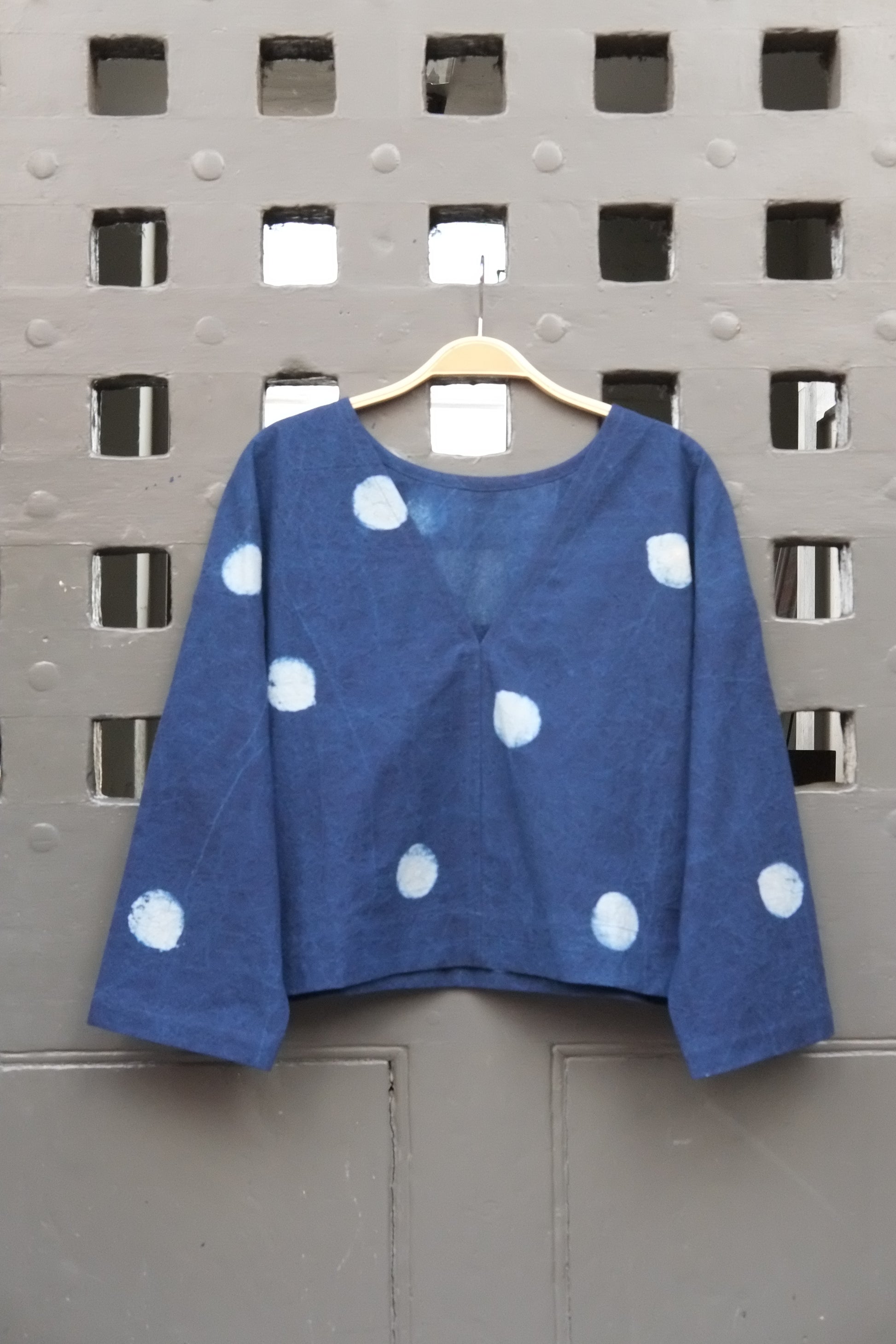 An indigo blue long-sleeve top with white polka dots hanging on a wooden hanger against a grey grid door