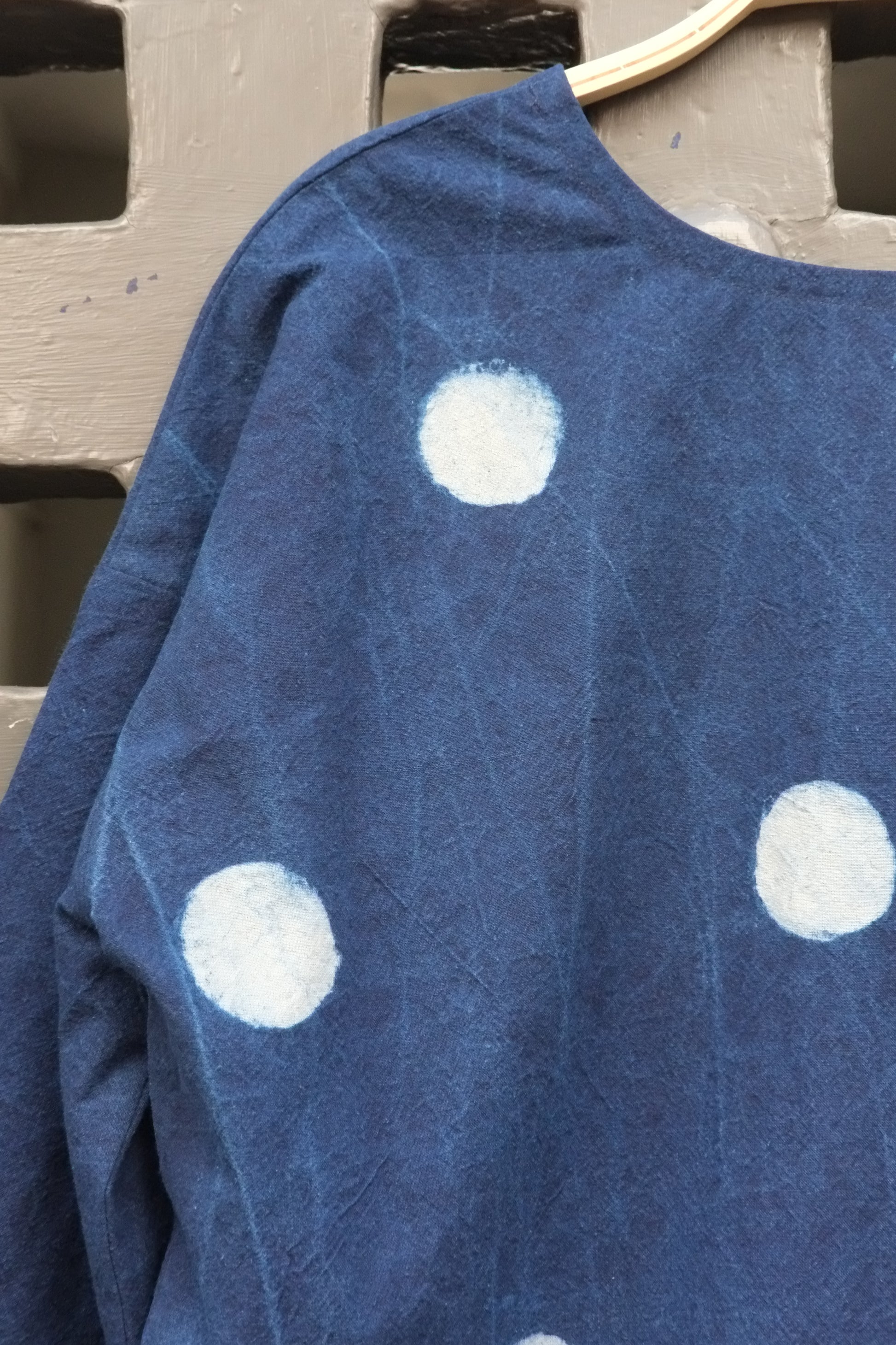 An indigo blue long-sleeve top with white polka dots hanging on a wooden hanger against a grey grid door