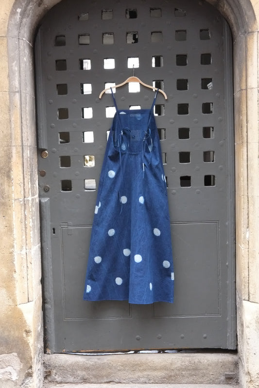 A blue sleeveless dress with white polka dots hangs from a wooden hanger on a dark metal gate with square cutouts, set against a stone wall background.