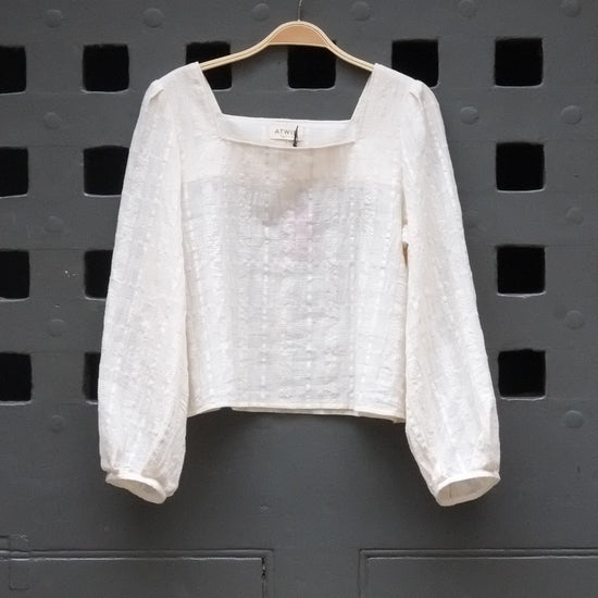 A white textured long-sleeve blouse with a square neckline, hanging on a wooden hanger against a dark gray perforated metal wall.