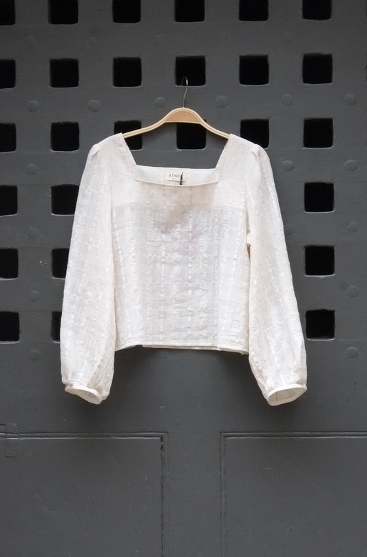 A white textured long-sleeve blouse with a square neckline, hanging on a wooden hanger against a dark gray perforated metal wall.