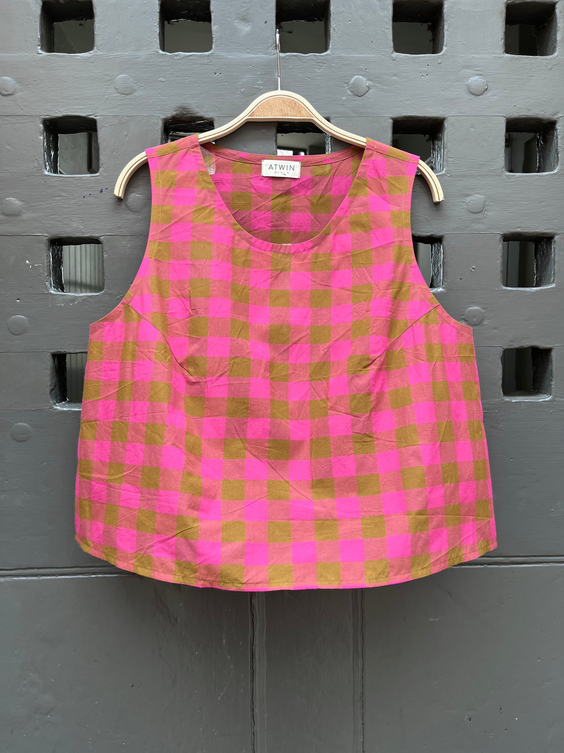 Fuchsia pink and khaki green checked cotton sleeveless tank top on a wooden hanger against a grey door