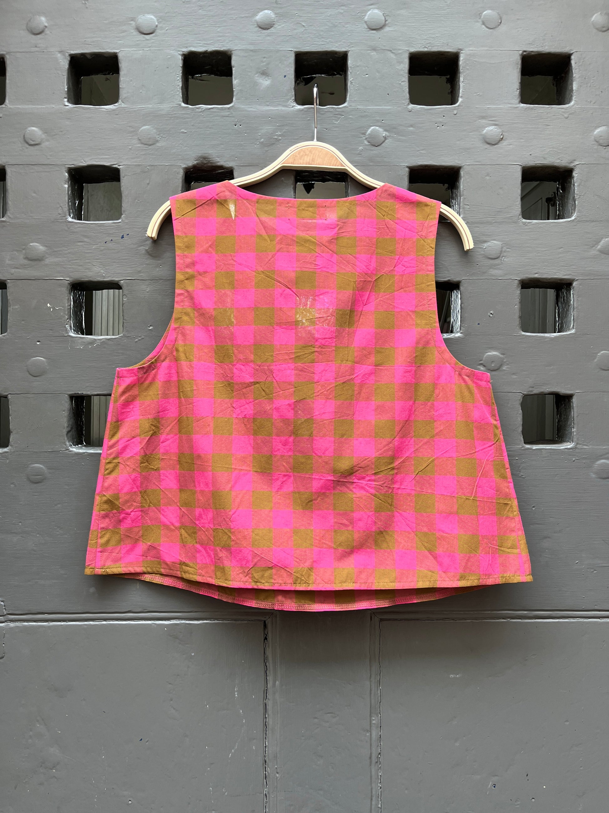 Fuchsia pink and khaki green checked cotton sleeveless tank top on a wooden hanger against a grey door