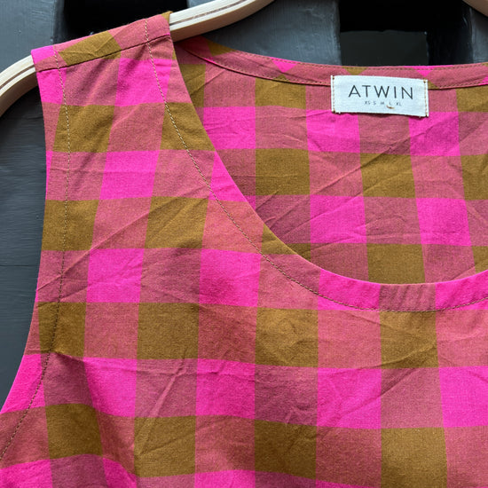 Fuchsia pink and khaki green checked cotton sleeveless tank top on a wooden hanger against a grey door