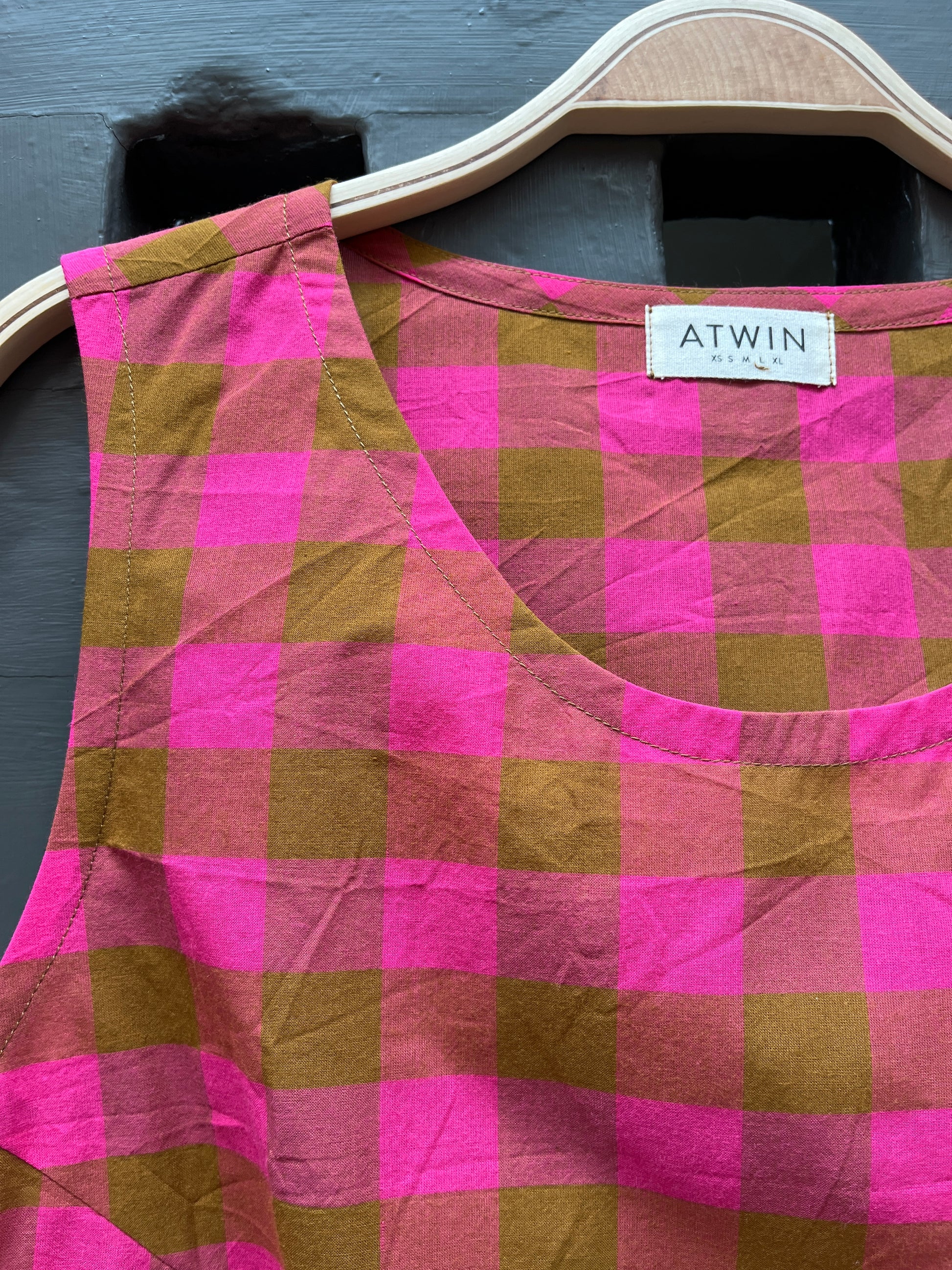 Fuchsia pink and khaki green checked cotton sleeveless tank top on a wooden hanger against a grey door