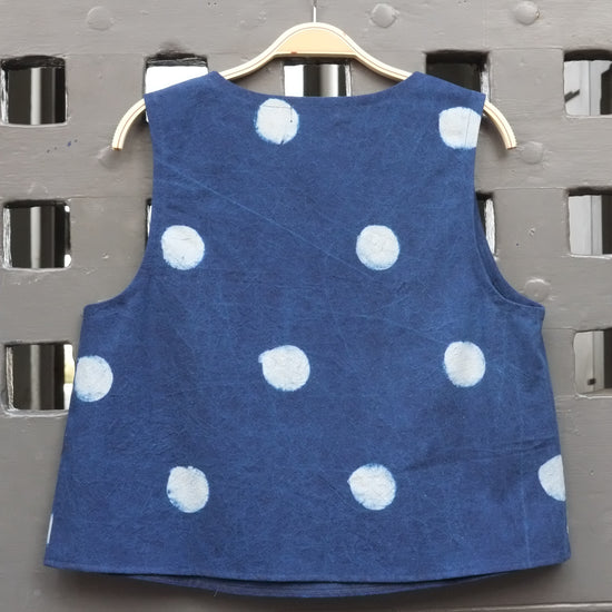 A sleeveless blue top with white polka dots hanging on a wooden hanger against a grey door.