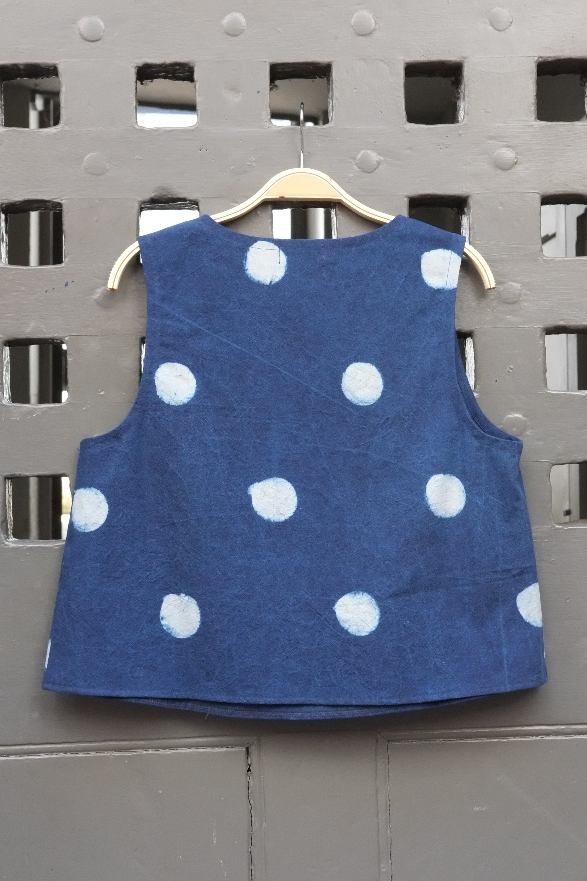 A sleeveless blue top with white polka dots hanging on a wooden hanger against a grey door.