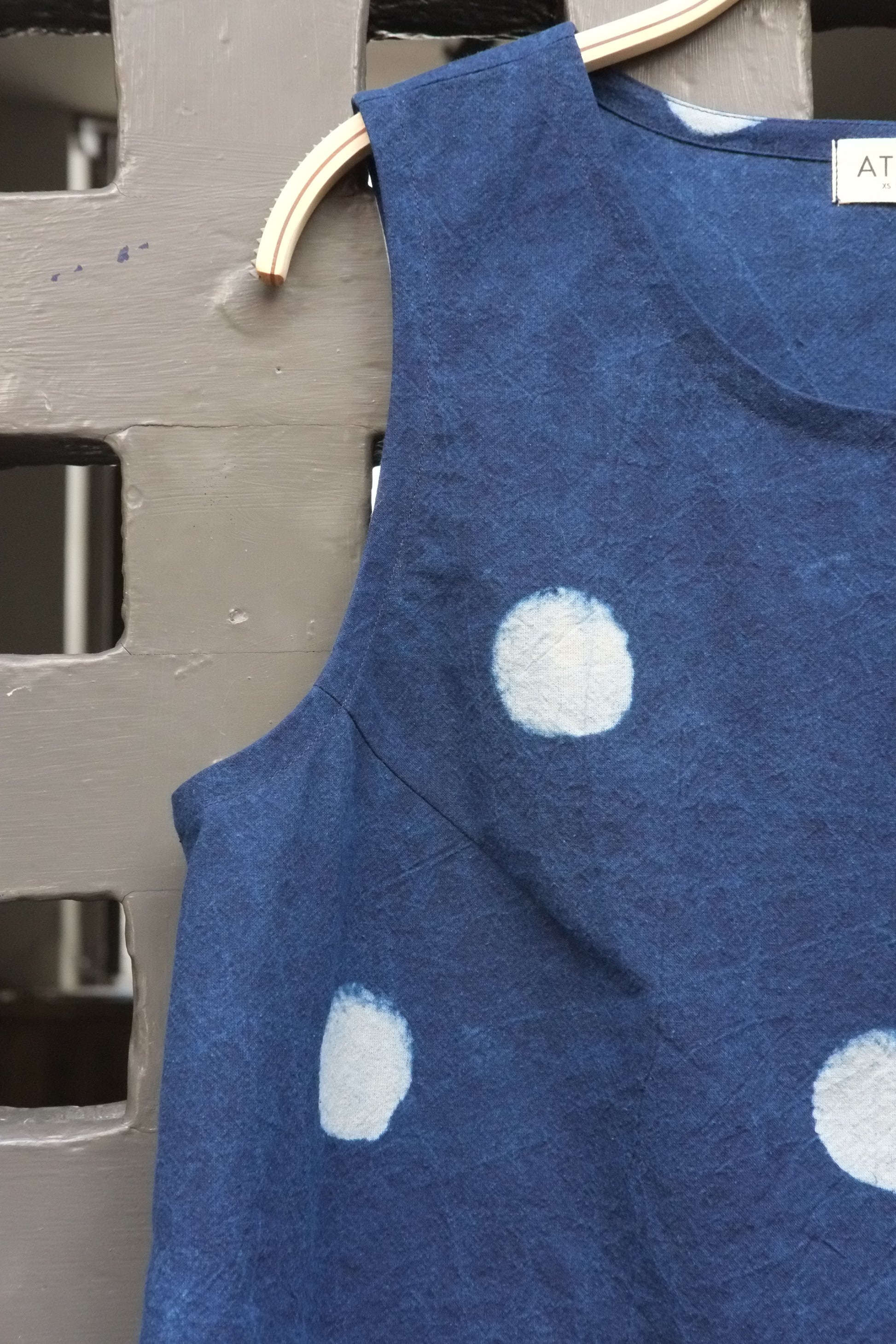 A sleeveless blue top with white polka dots hanging on a wooden hanger against a grey door.