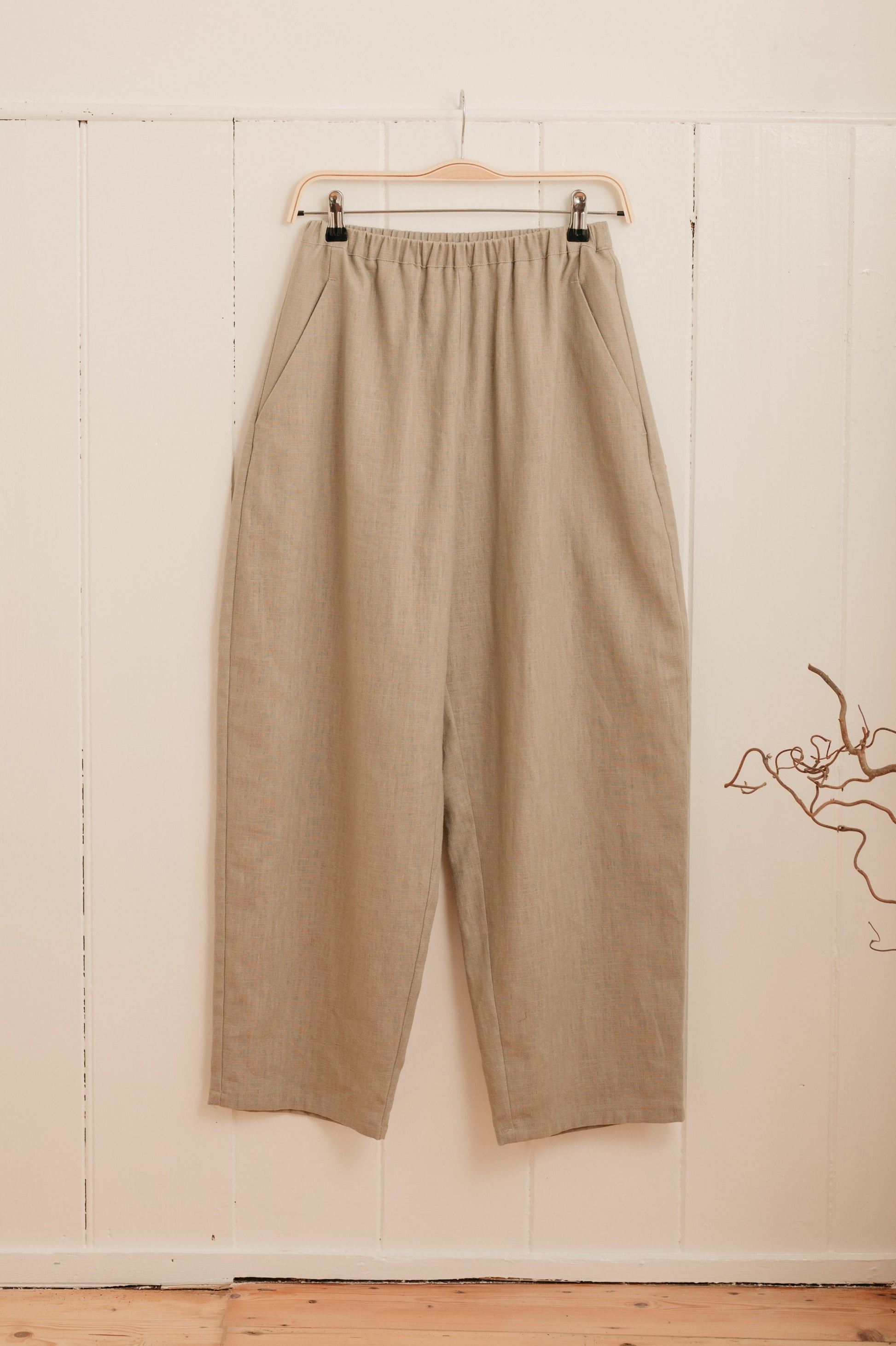 A pair of beige barrel leg trousers hanging on a hanger against a white wall, with a slim elasticated waistband and a high-waisted cut.