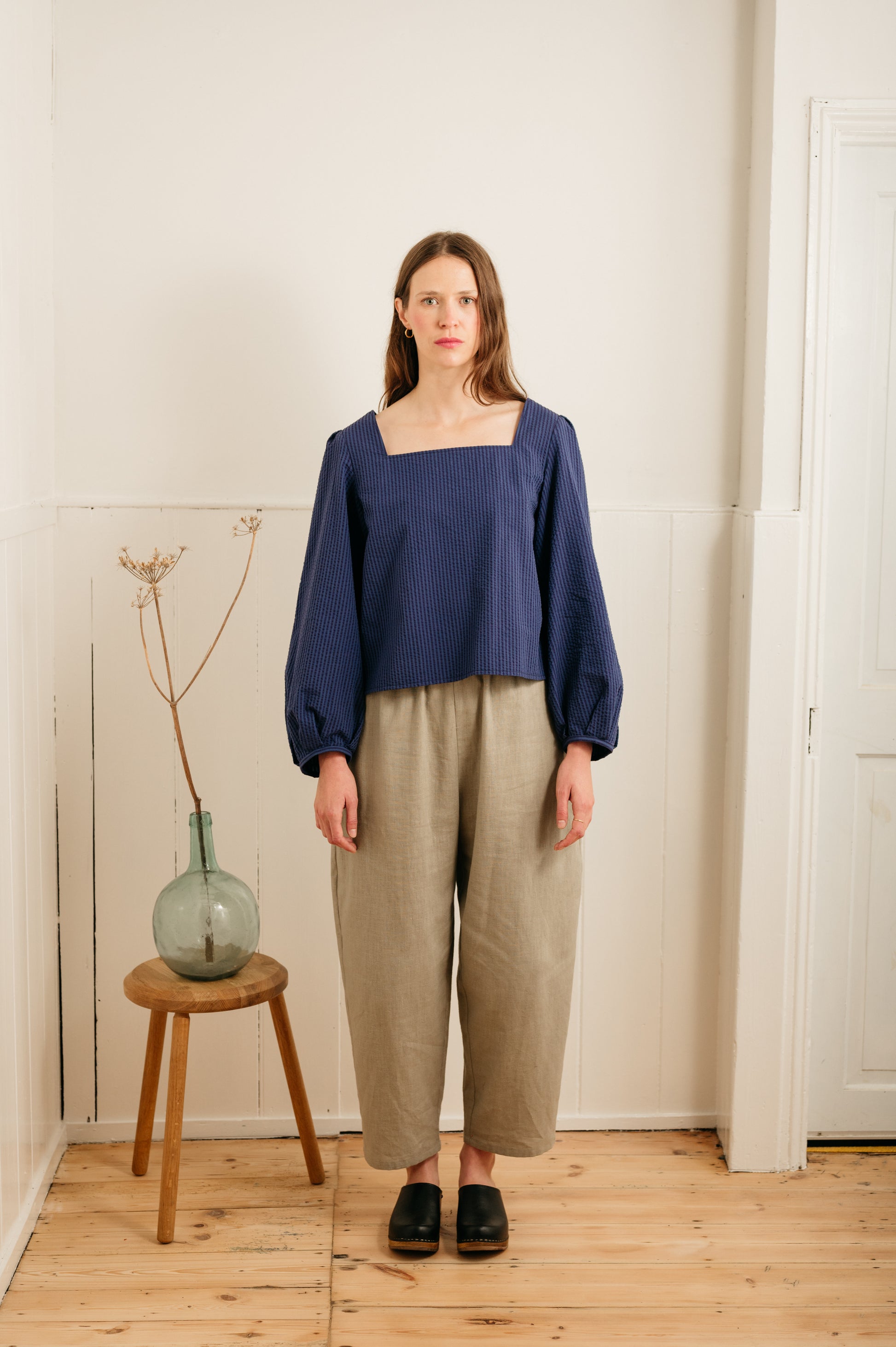 woman wearing atwin the label grain barrel leg trousers with a blue top against a wall
