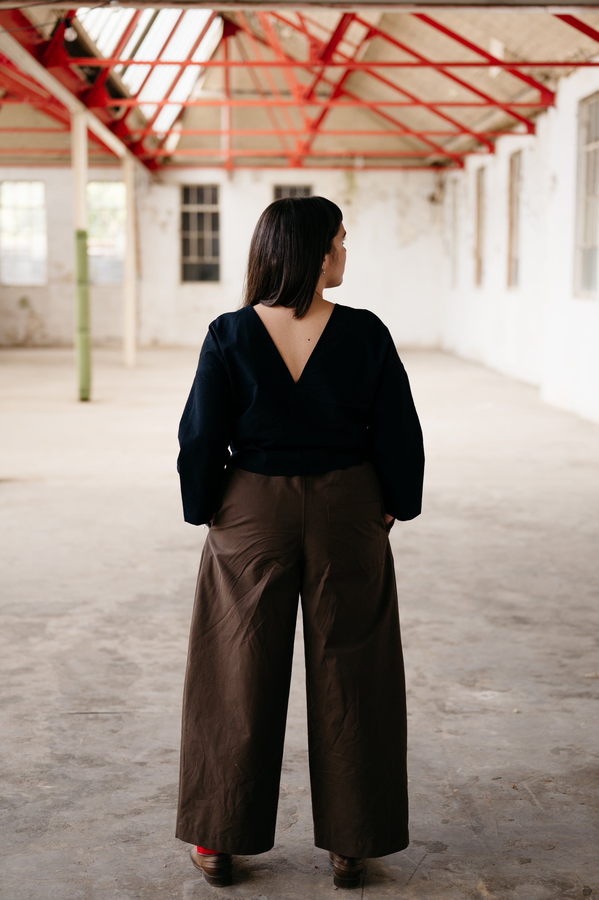 Person wearing a black top and brown wide-leg pants in an empty warehouse.
