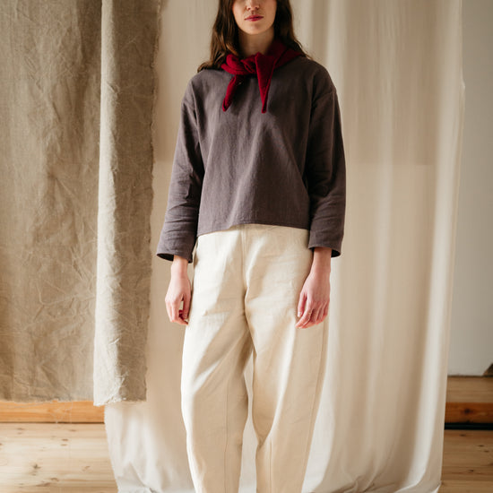 A woman standing in an ecru denim barrel leg trouser with a relaxed fit, featuring a slim elasticated waistband and high-waisted cut. She is also wearing a gray top with a round neckline and a red ribbon tie at the neck.