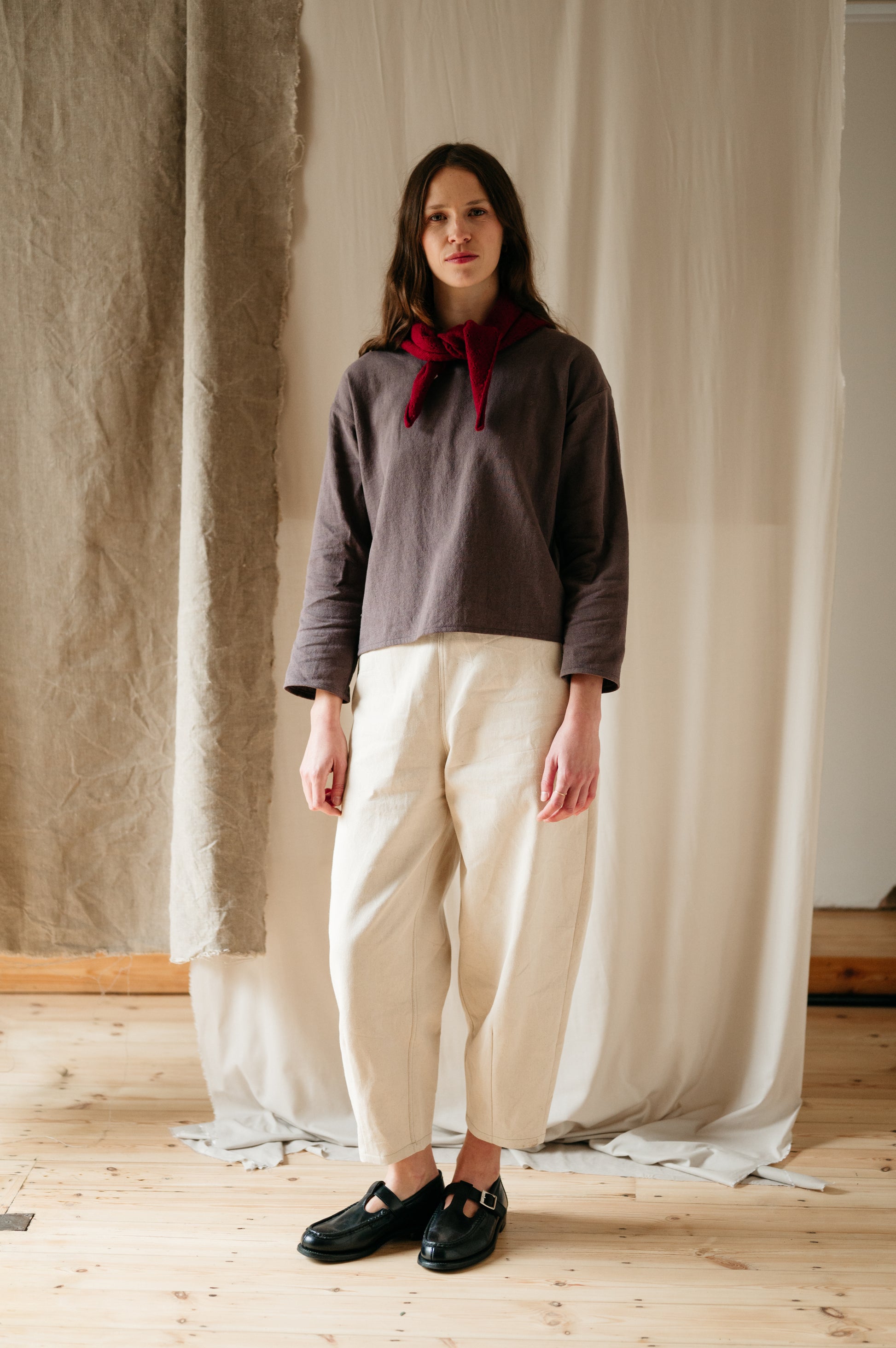 A woman standing in an ecru denim barrel leg trouser with a relaxed fit, featuring a slim elasticated waistband and high-waisted cut. She is also wearing a gray top with a round neckline and a red ribbon tie at the neck.
