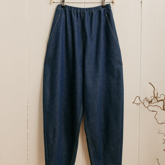 A pair of indigo denim barrel leg trousers with an elasticated waistband hanging on a hanger against a white wooden background.