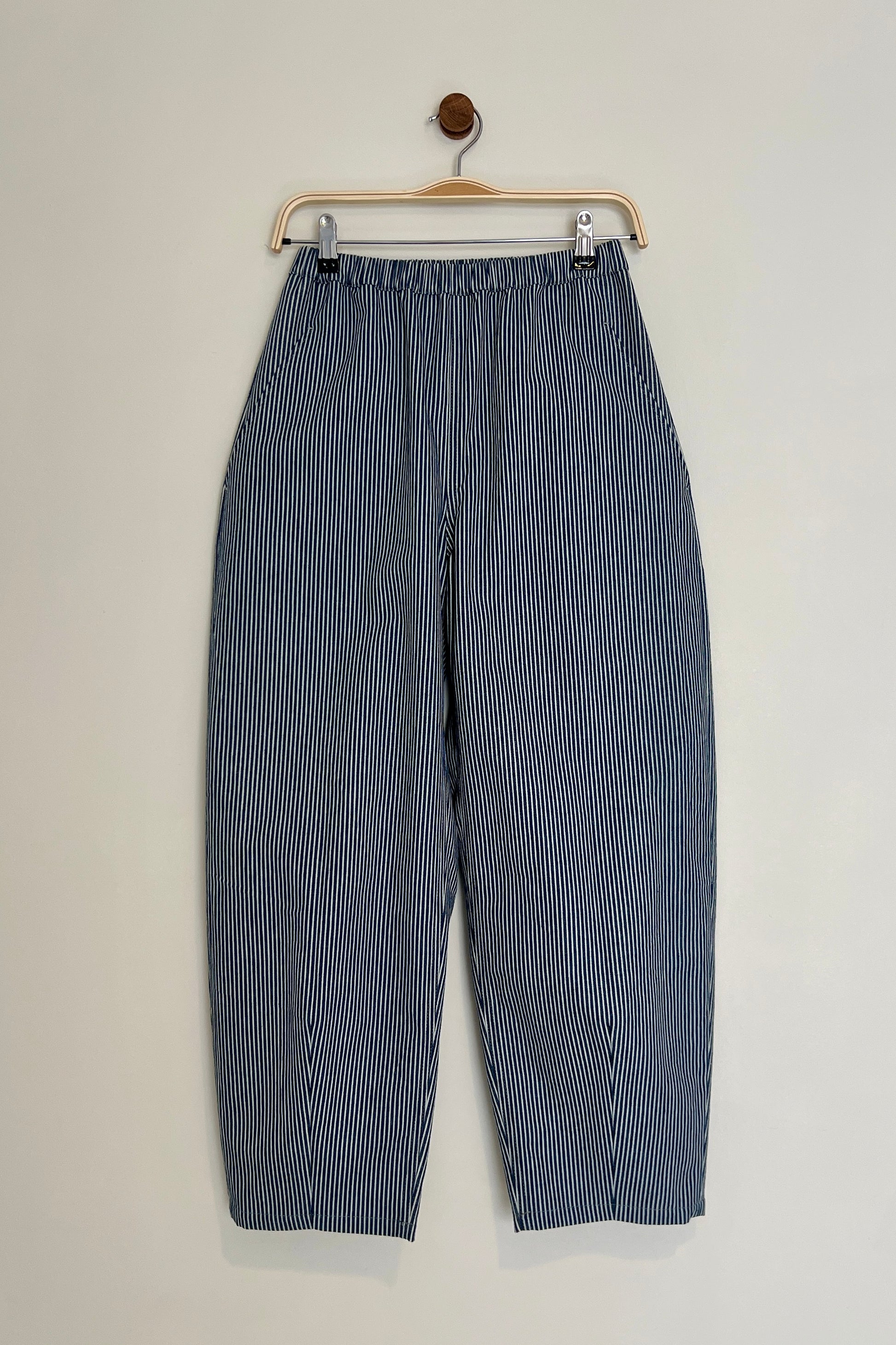 Blue and white striped barrel leg trousers hanging on a wooden hanger against a light gray background