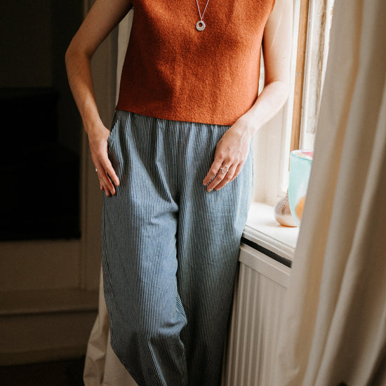 Person wearing a rust-coloured sleeveless top and blue striped jeans standing by a window.
