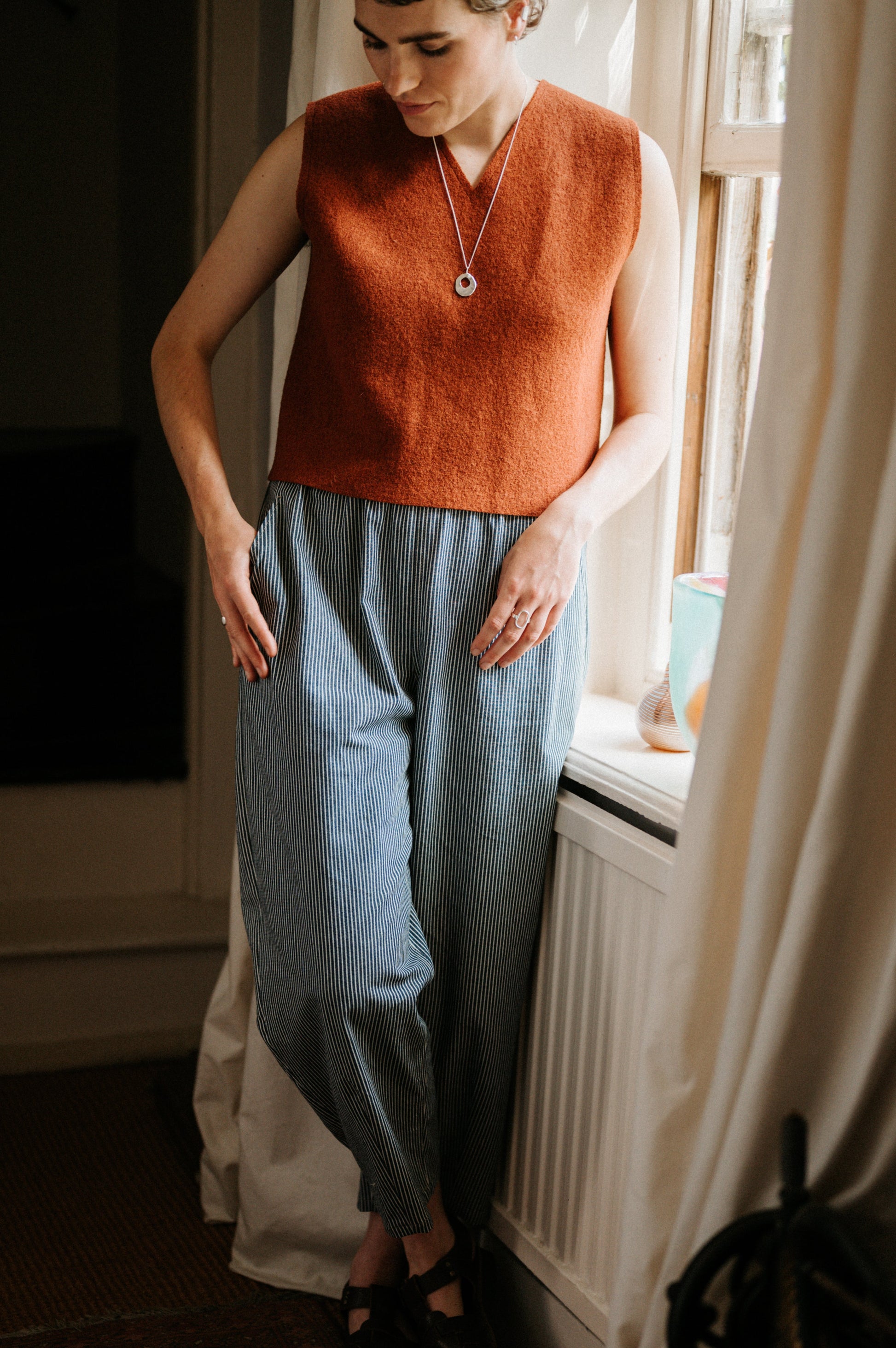 Person wearing a rust-coloured sleeveless top and blue striped jeans standing by a window.