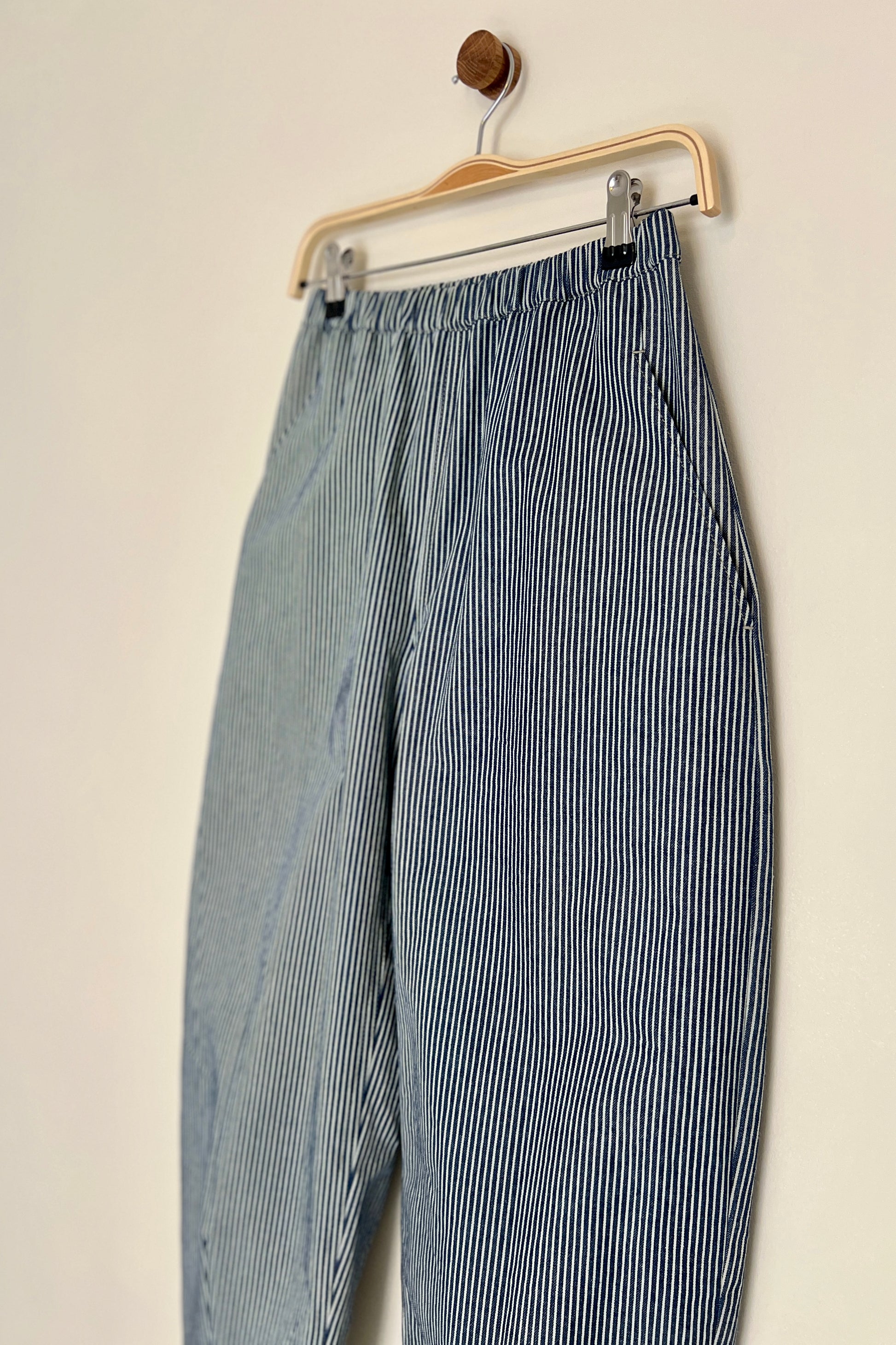 Blue and white striped denim trousers hanging on a wooden hanger against a beige wall.