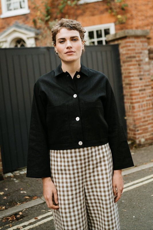 Person wearing a black jacket and checkered pants standing in front of a brick building.