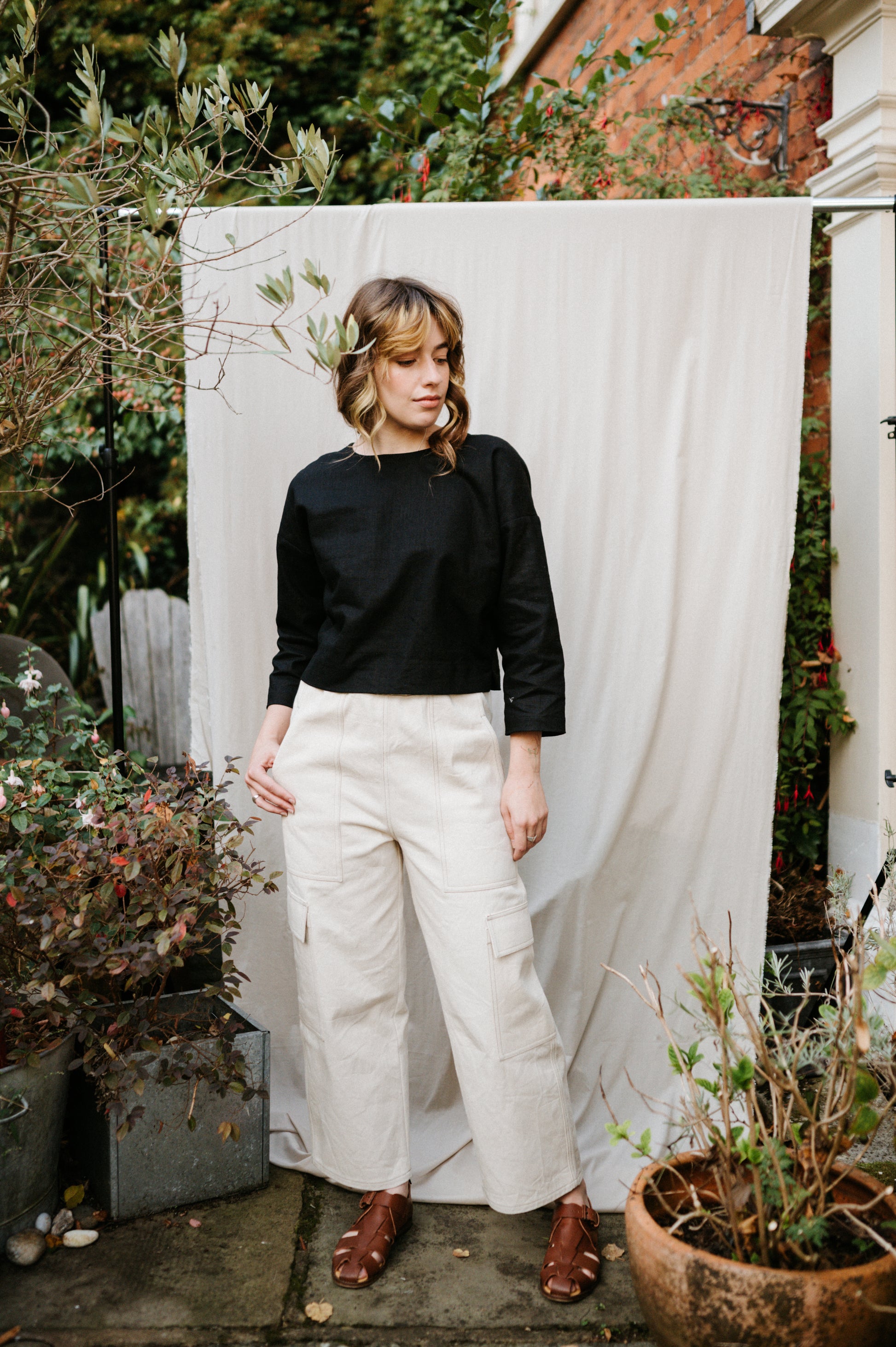 Person wearing a black top and ecru cargo jeans standing in front of a white curtain with plants around.