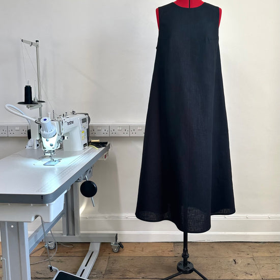 A black sleeveless, high neck dress with a deep v-back on a mannequin in a sewing studio setting, indicating a fitted garment made from ramie linen.