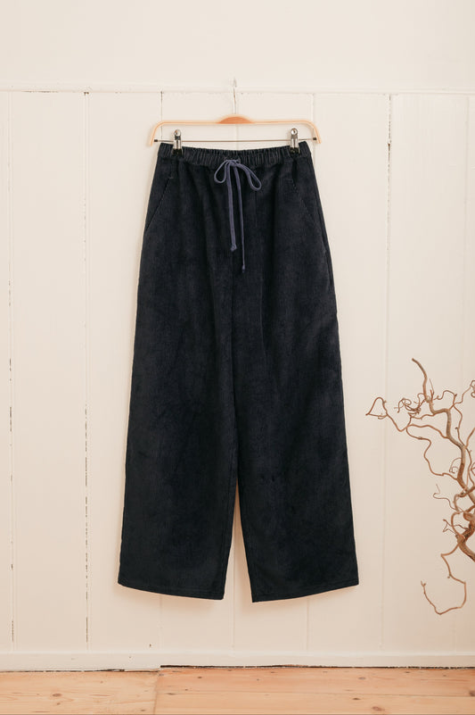 A pair of navy corduroy trousers with a drawstring waistband hanging on a hanger against a white background, with a subtle blue cord drawstring detail.