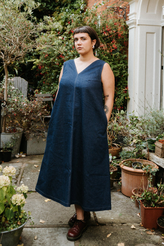 Woman in a blue denim dress standing in a garden with plants and a house in the background
