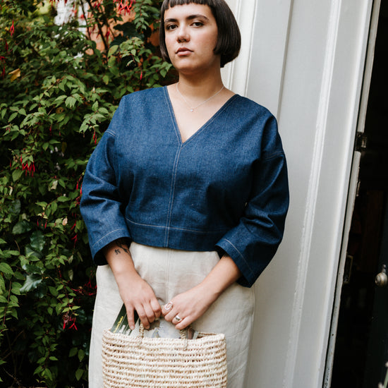 Woman in an indigo denim top and beige pants holding a woven bag, standing outdoors.