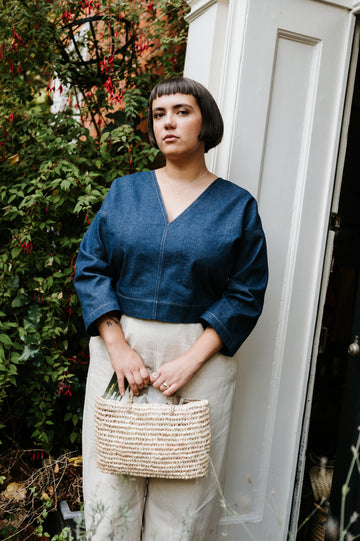 Woman in an indigo denim top and beige pants holding a woven bag, standing outdoors.