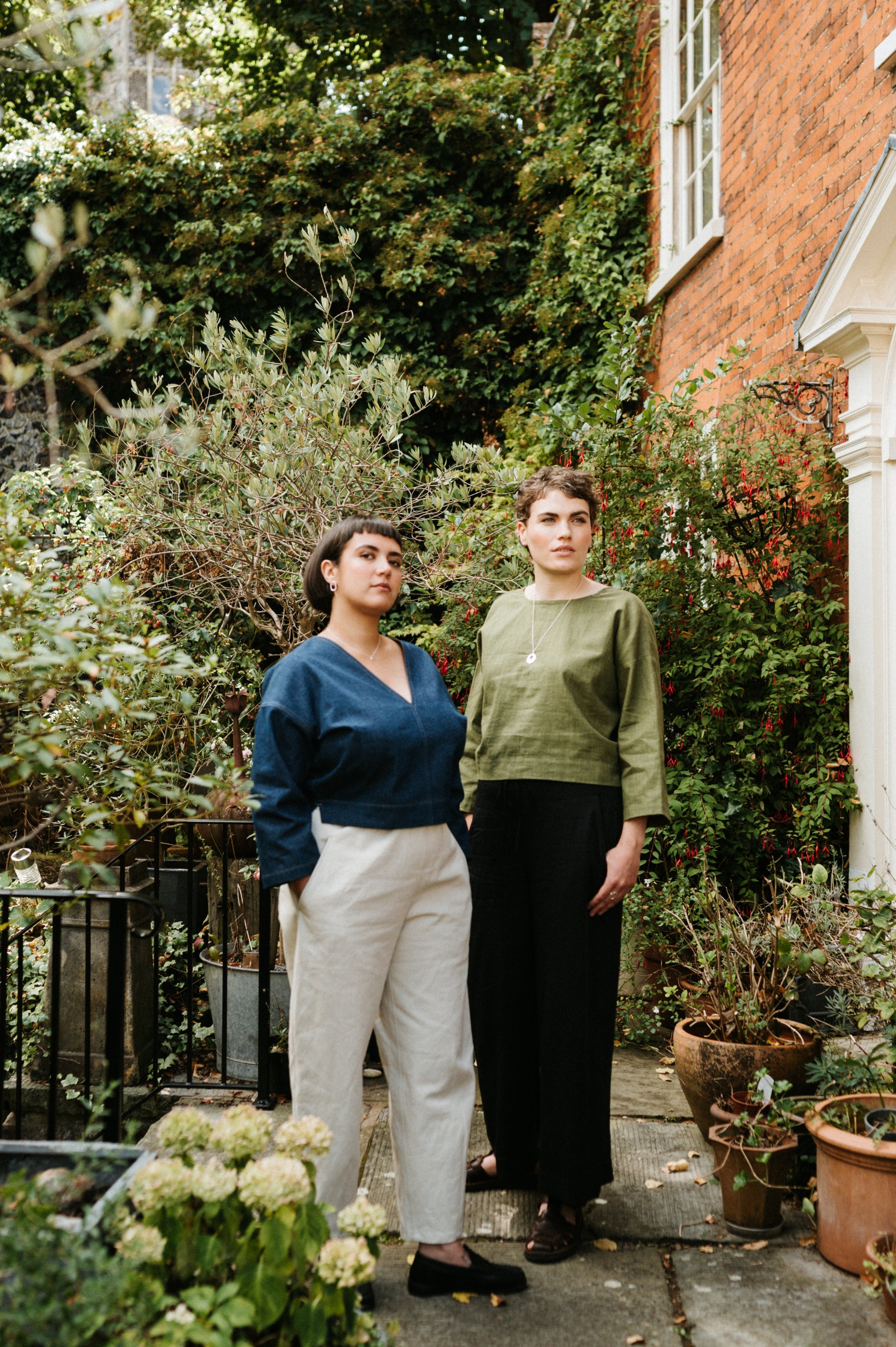Two women standing in a garden with greenery and a house in the background