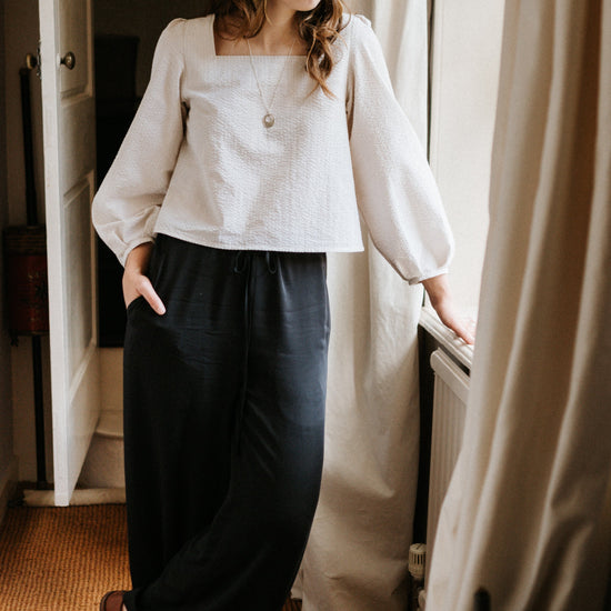Woman in a white blouse and dark pants standing in a room with a window and curtain.