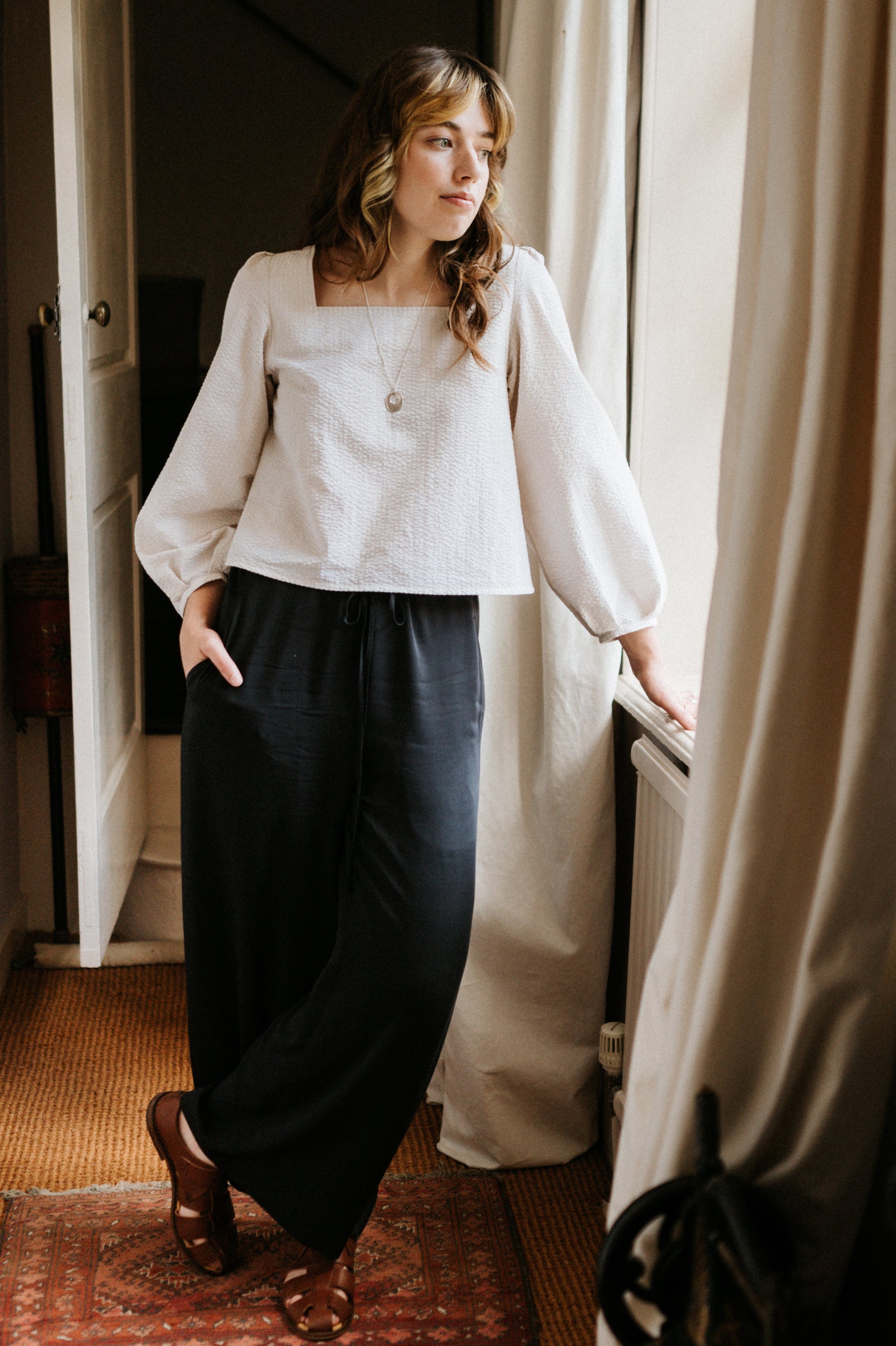 Woman in a white blouse and dark pants standing in a room with a window and curtain.