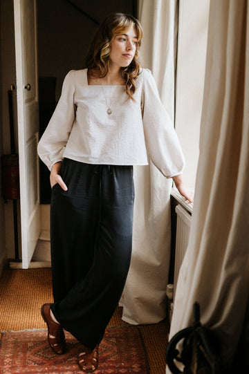 Woman in a white blouse and dark pants standing in a room with a window and curtain.