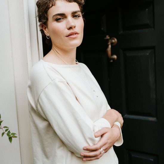 Woman standing in a doorway wearing a ecru smock top and blue jeans.