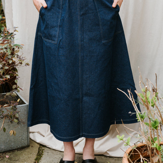 Person wearing a blue denim utility skirt with plants and a white curtain in the background