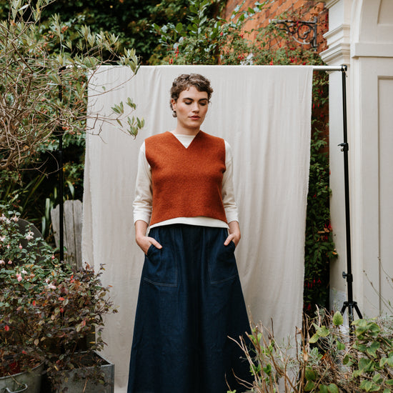 Person wearing a rust-colored sweater and dark blue skirt standing outdoors with plants and a white backdrop.
