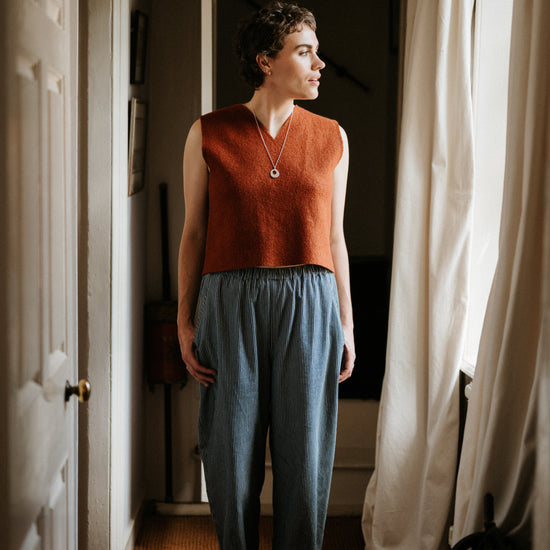 Woman standing in a room with a window and curtain, wearing a rust-colored sleeveless top and blue pants.