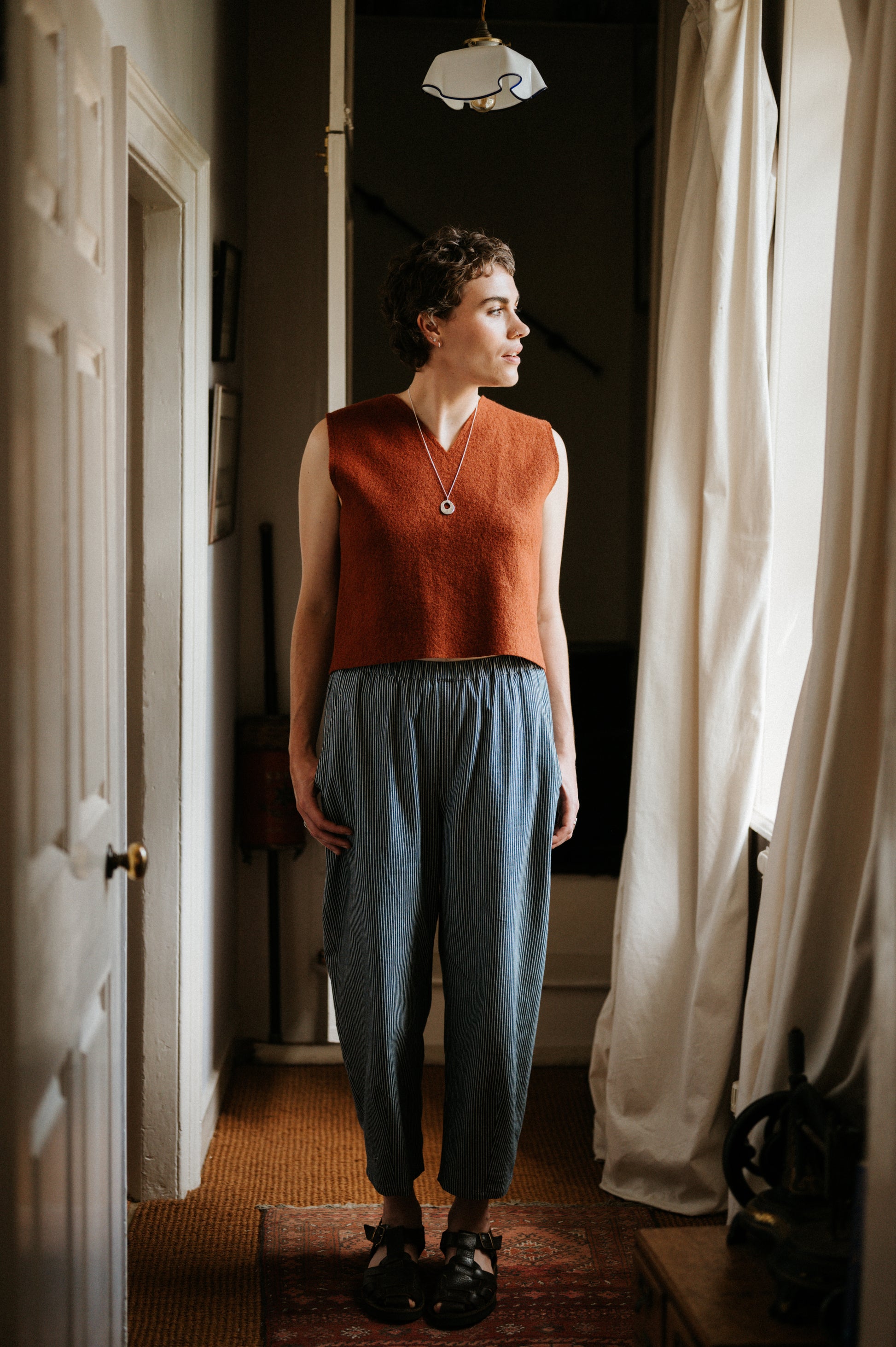 Woman standing in a room with a window and curtain, wearing a rust-colored sleeveless top and blue pants.