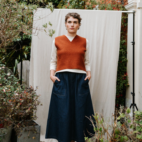 Person wearing a rust-coloured boiled wool vest and dark blue skirt standing in a garden.