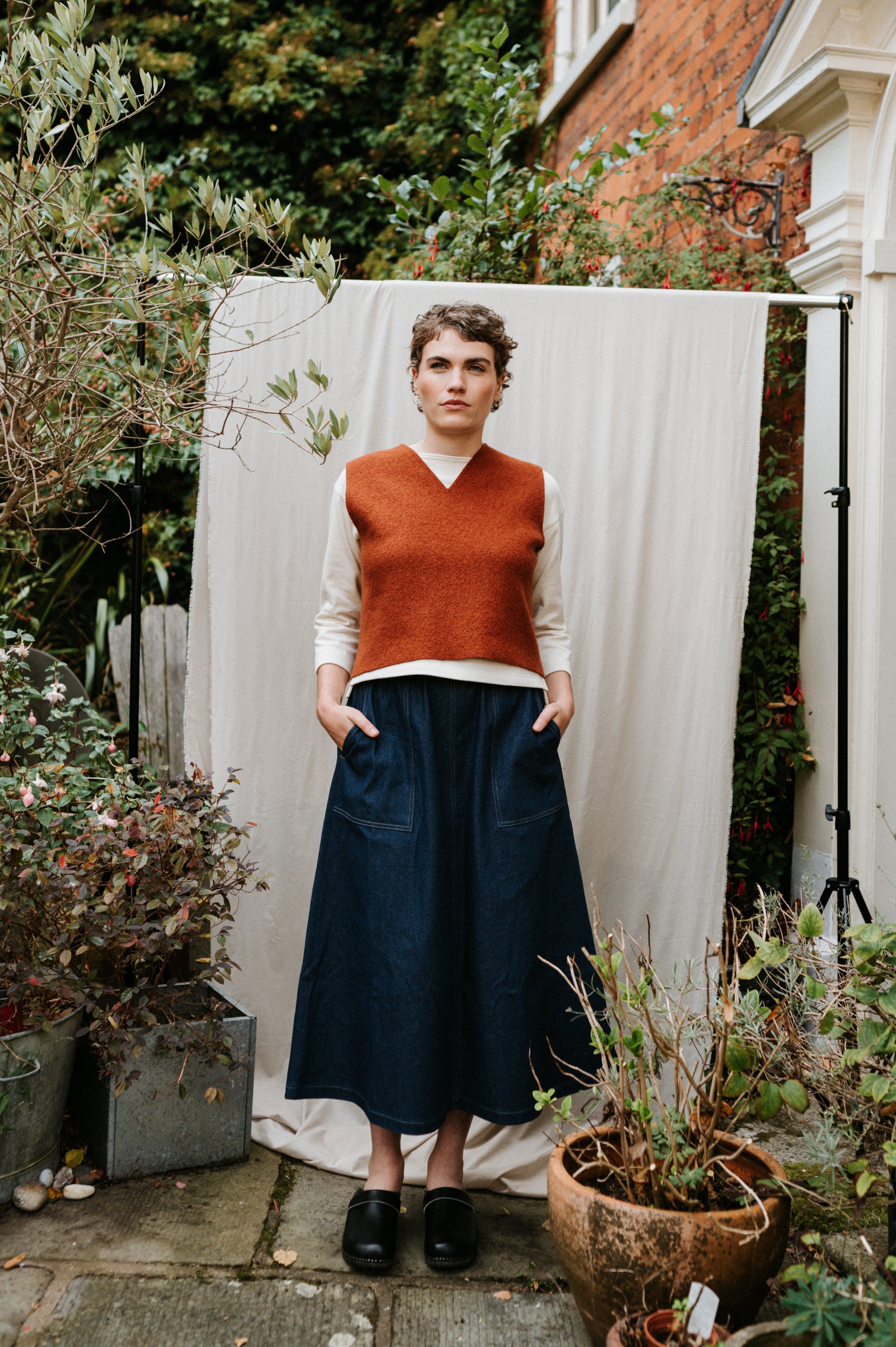 Person wearing a rust-coloured boiled wool vest and dark blue skirt standing in a garden.